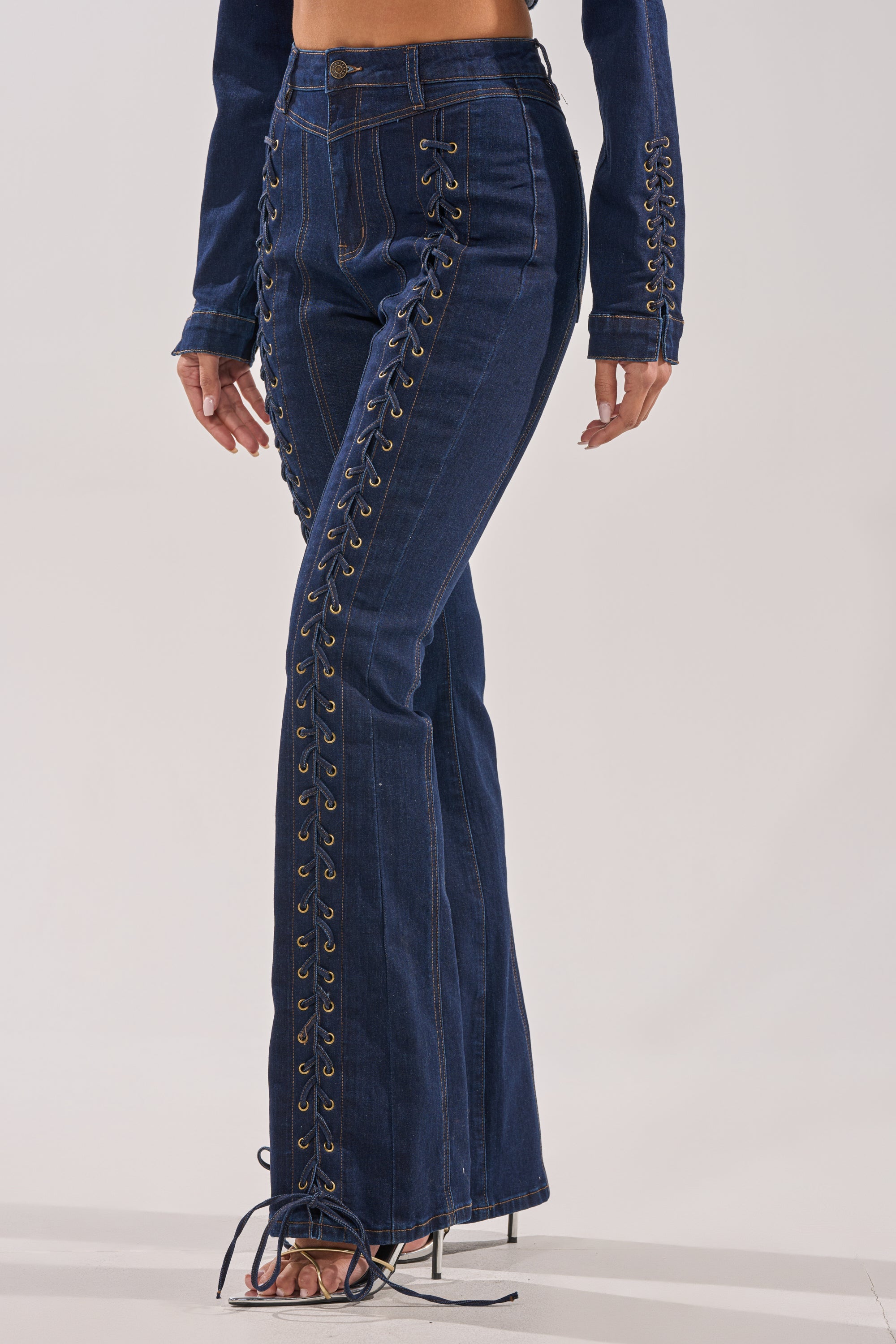 A person wears the THATS ON ME FLARE DENIM PANT, featuring dark blue high-rise flare jeans with front lace-up details, paired with heeled sandals. The image is cropped from the shoulders down. 0