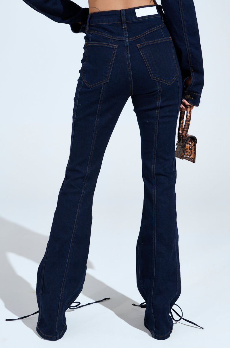 A person wearing the THATS ON ME FLARE DENIM PANT in dark blue with a high-rise fit and matching jacket, holding a small brown handbag. The jeans feature prominent stitching and a waistband label against a plain white background.