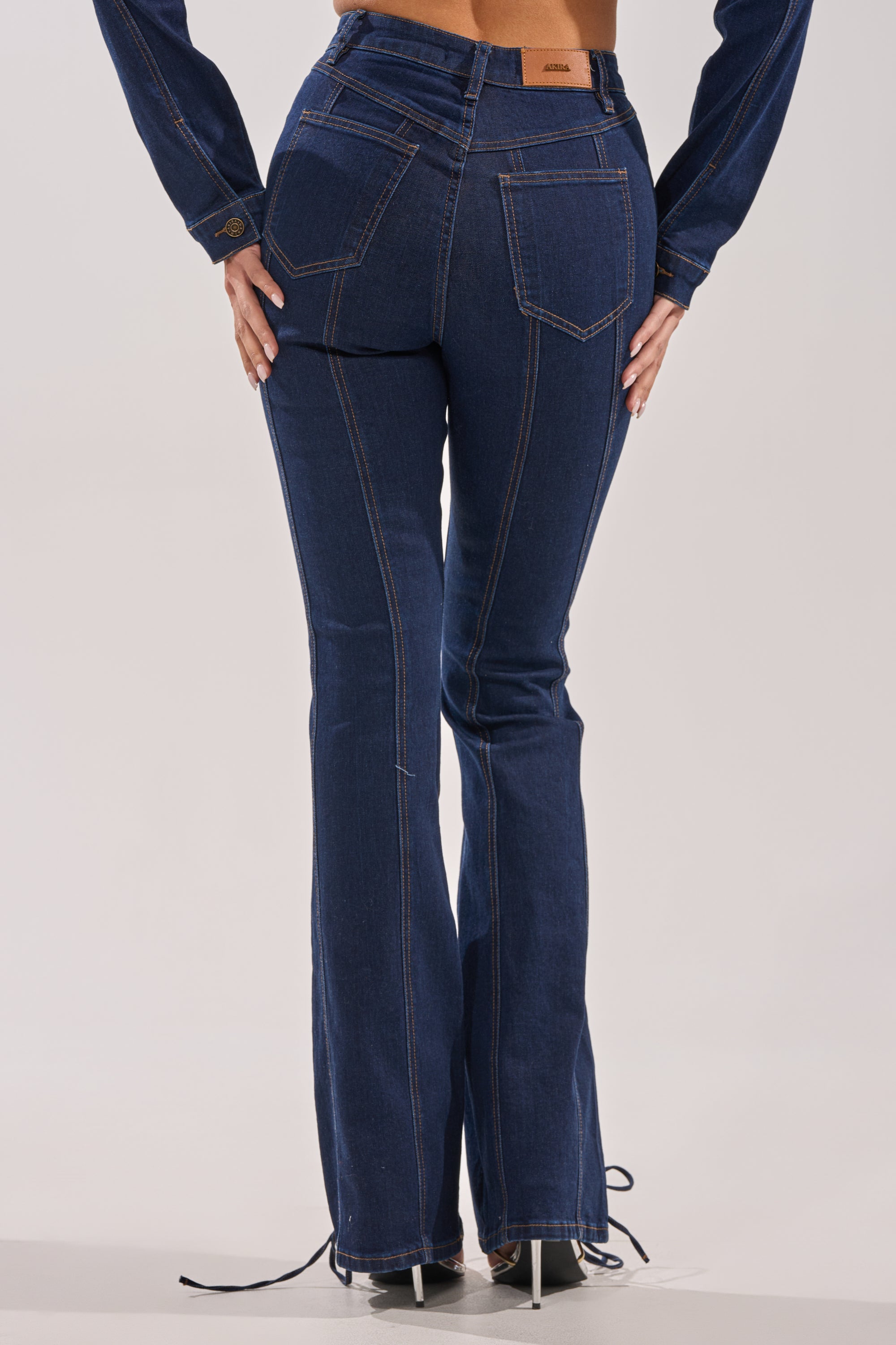 A person, seen from behind, wears the THATS ON ME FLARE DENIM PANT in dark blue. The high-rise jeans feature visible stitching and a slight flare at the bottom, nearly covering their high-heeled shoes. 3