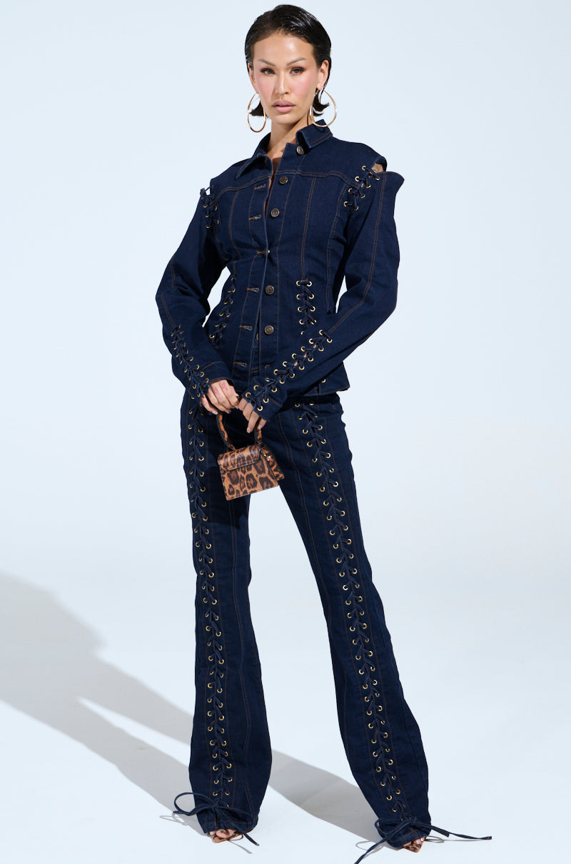 A person stands confidently wearing a fitted dark denim jacket and THATS ON ME FLARE DENIM PANT with lace-up details. They hold a small leopard print bag and sport large hoop earrings against a plain light background.