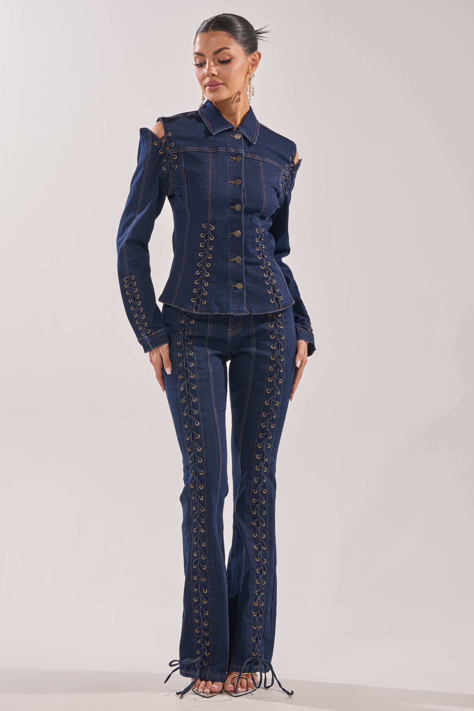 A woman stands barefoot, hair tied back, wearing the THATS ON ME FLARE DENIM PANT set featuring a fitted, dark flare design with cut-out shoulders, button details, and laces along the arms, legs, and front seams. 4