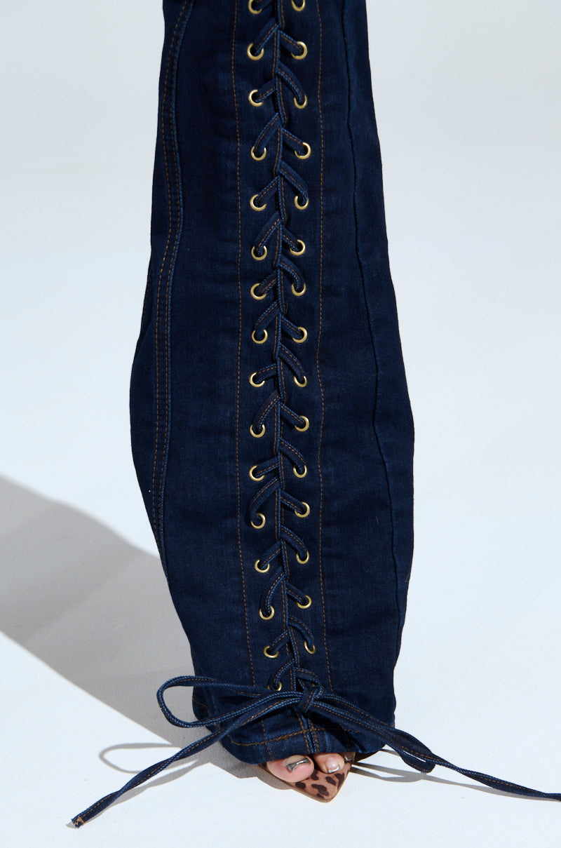 The THATS ON ME FLARE DENIM PANT showcases a high-rise, dark denim design. Gold eyelets and blue laces run vertically from ankle to knee, ending in a bow. A close-up highlights the foot subtly resting on a light, smooth surface.