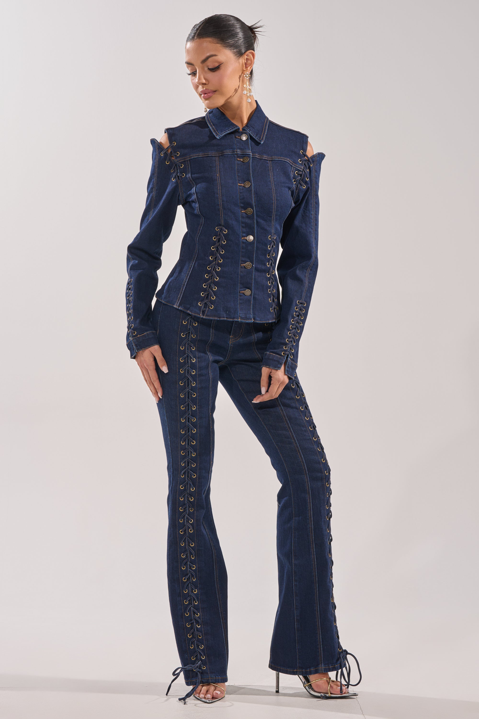 A woman models the THATS ON ME FLARE DENIM PANT, paired with a dark blue denim jacket. The high-rise pants feature lace-up seam detailing. She poses with her hair pulled back, one hand on her hip, and looks downward. 5