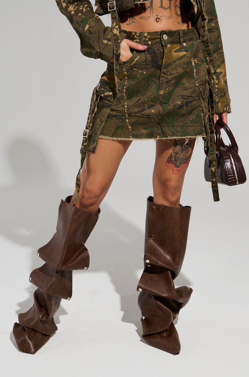 A person is dressed in a camouflage pattern jacket paired with the Running Free Mini Skirt and a coordinating crop top. They complete the ensemble with high, layered brown boots and carry a small brown handbag. Their thigh displays a tattoo against a neutral background.