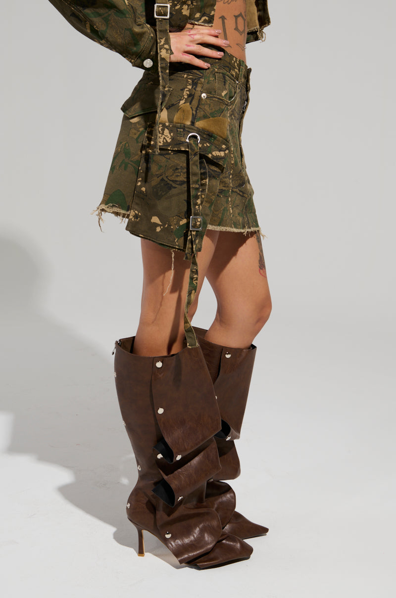 A person is dressed in a camouflage-patterned ensemble, including the RUNNING FREE MINI SKIRT and a matching jacket. They pair this look with large, over-the-knee brown leather boots adorned with oversized straps and buckles. Their midriff is exposed as they pose confidently against a plain backdrop.