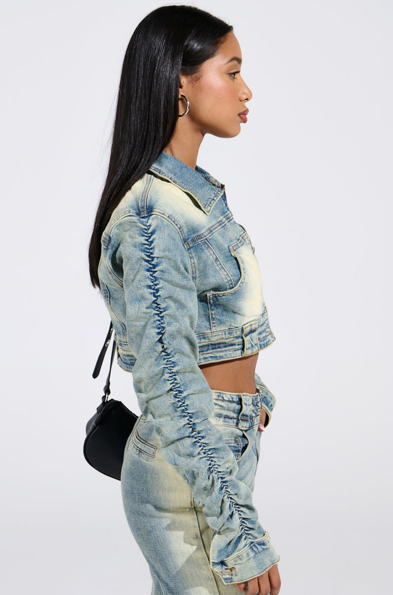 A person with long black hair is wearing an UP ALL NIGHT DENIM JACKET with ruched sleeves and matching jeans. They have hoop earrings, a small black shoulder bag, and are turned to the side against a plain background.
