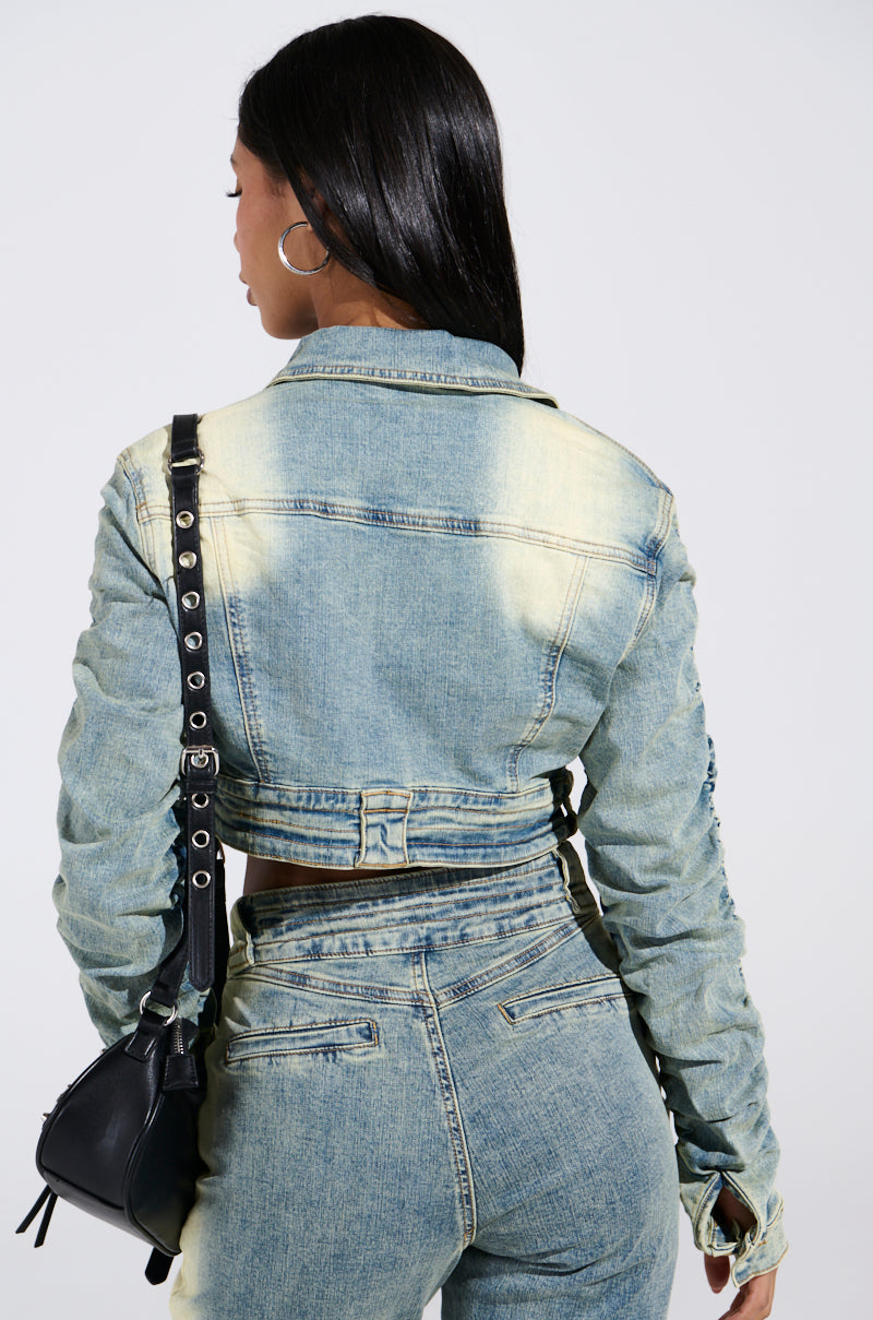 A person with long black hair, hoop earrings, and a black shoulder bag is facing away from the camera in a cropped denim jacket and jeans. The plain, light backdrop highlights their style featuring the UP ALL NIGHT DENIM JACKET.