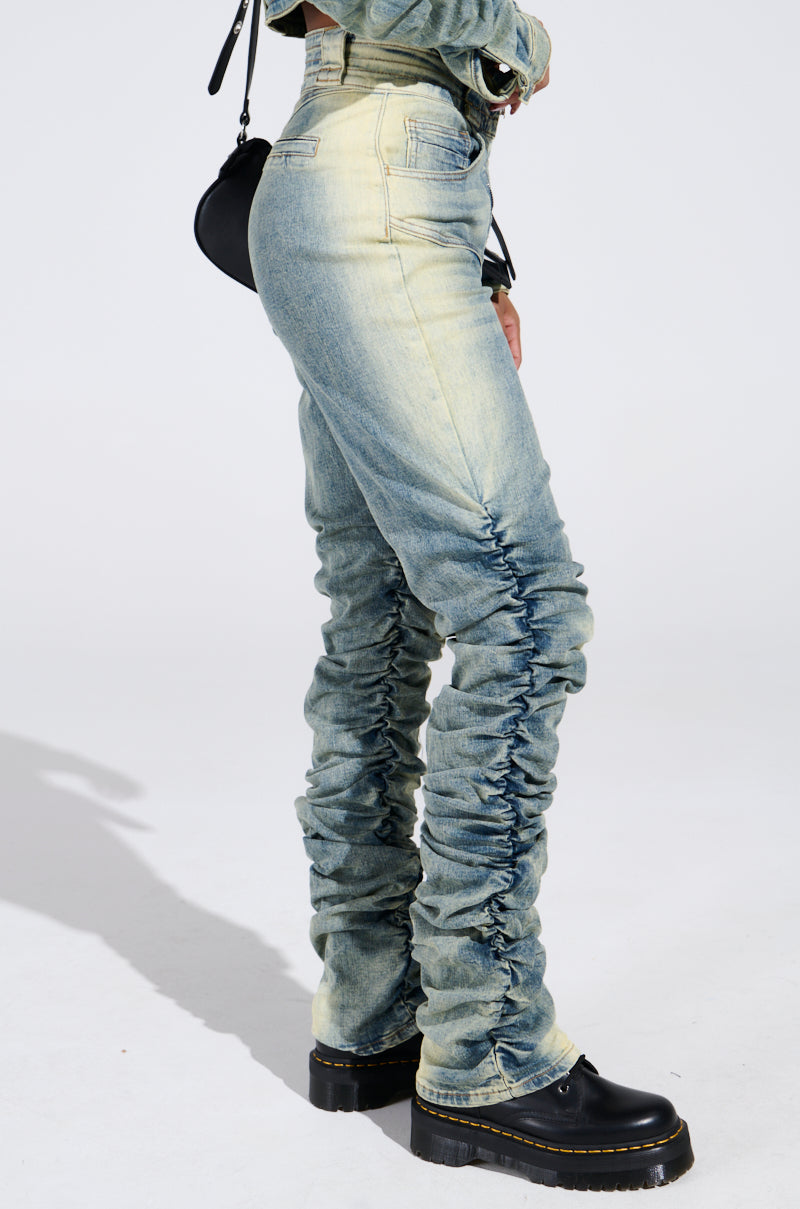 The person is wearing UP ALL NIGHT DENIM PANT in light blue, ruched high-rise style with a faded wash paired with black platform shoes and a small black handbag, all against a crisp white background that highlights their fashionable attire.