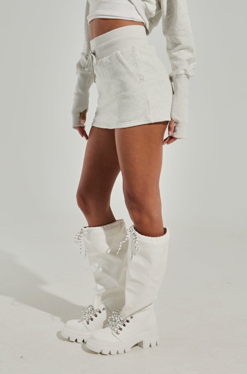 A person stands against a neutral background, adorned in a light gray cropped sweater and the STINGY WITH MY TIME MINI SKIRT. Complementing the ensemble are knee-high white boots with lace-up details and chunky soles. Their face is not shown in the image.