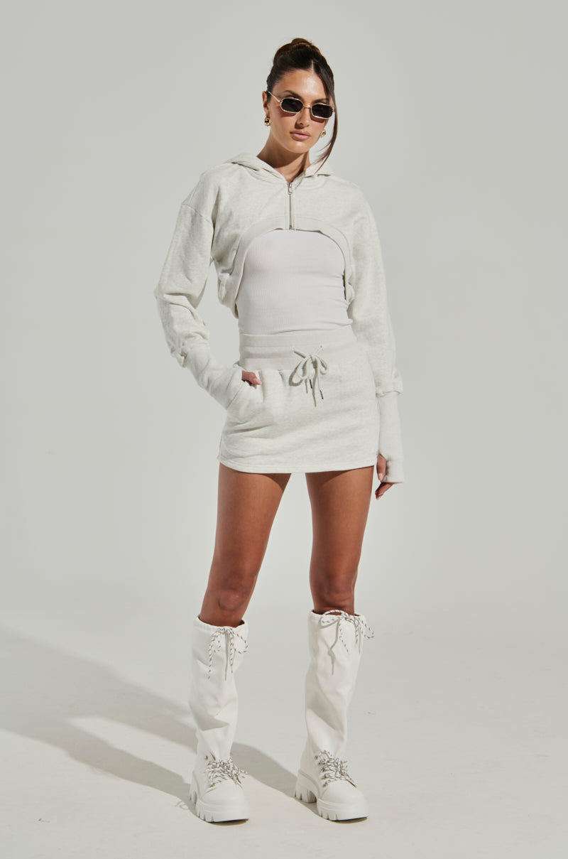 A woman stands confidently against a plain background. She wears a white zip-up hoodie and the STINGY WITH MY TIME MINI SKIRT, made from a stretchy cotton blend, paired with high boots. She accessorizes with sunglasses and has her hair pulled back. Her outfit gives a casual yet stylish look.