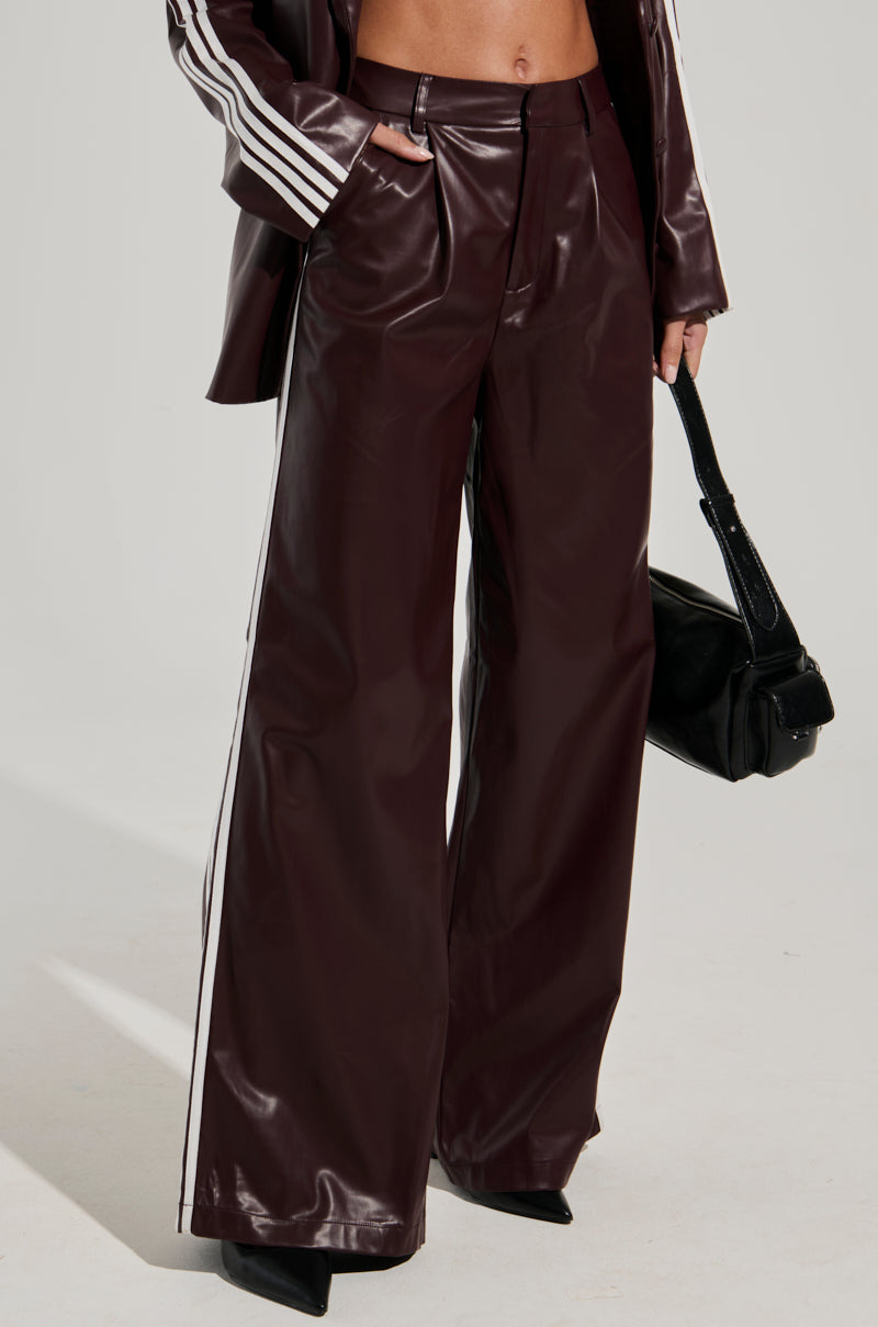 A person is wearing a stylish outfit consisting of the DAYTIME LOVIN TROUSER IN BURGUNDY and a matching jacket with white side seam stripes. They hold a black handbag in one hand, and black pointed-toe high heels are partially visible at the bottom.
