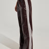 A person stands modeling the DAYTIME LOVIN TROUSER IN BURGUNDY, featuring dark brown, shiny faux leather with white side seam stripes. The high-rise wide-leg pants are loose-fitting, and the model's hand rests on their hip. The background is plain and light-colored.