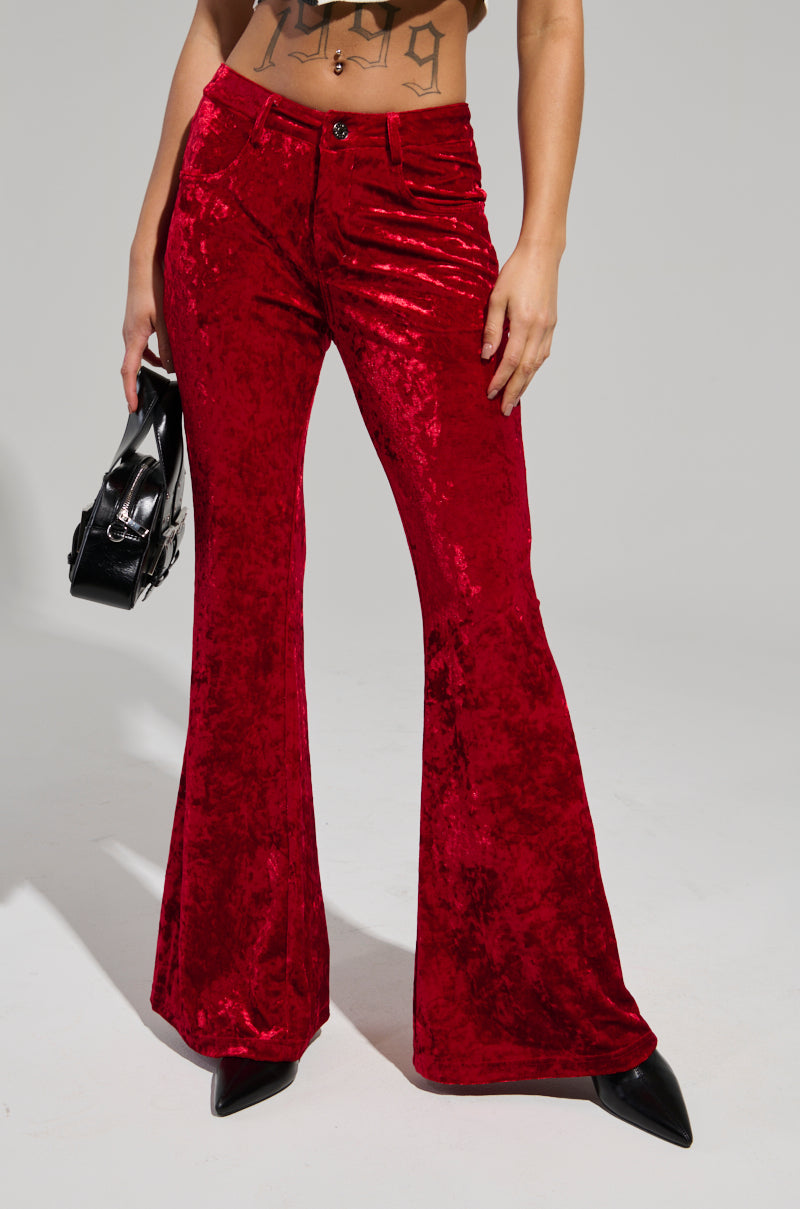 A person wearing the MEET ME HALF WAY TROUSER in a crushed velvet, high-rise fit with a flared silhouette is holding a black pointed shoe and handbag. The top part of their body is not visible, and the pants' shiny texture stands out against the plain gray background.