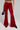 A person wearing the MEET ME HALF WAY TROUSER in a crushed velvet, high-rise fit with a flared silhouette is holding a black pointed shoe and handbag. The top part of their body is not visible, and the pants' shiny texture stands out against the plain gray background.