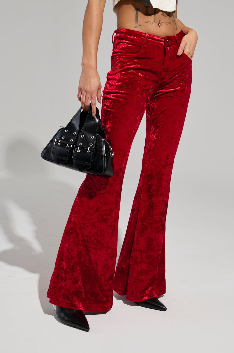 A person wearing the MEET ME HALF WAY TROUSER in red crushed velvet, featuring a flared silhouette, paired with black pointed shoes, is holding a black handbag adorned with metal accents against a plain grey background.