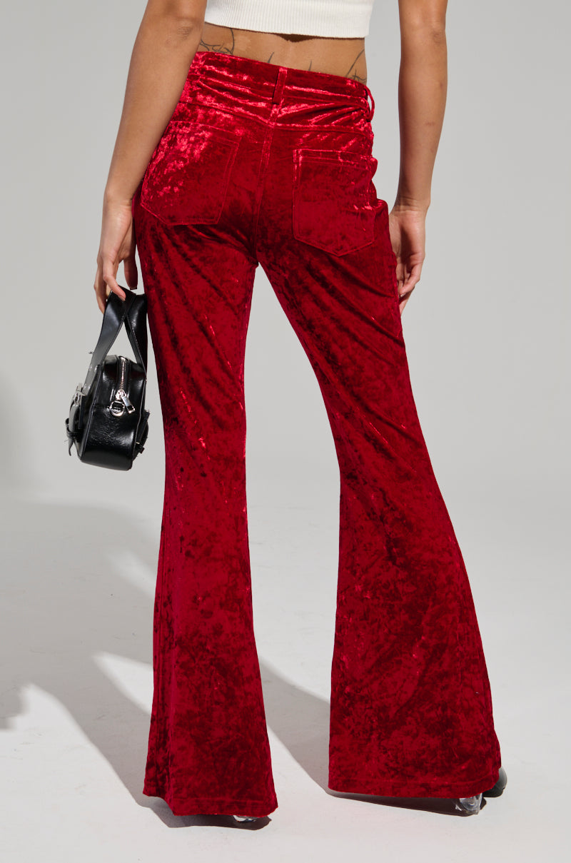 A person wearing MEET ME HALF WAY TROUSER in red crushed velvet and a beige cropped top carries a black handbag. The image captures the person from behind, highlighting the high-rise fit and texture of the trousers.