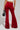 A person wearing MEET ME HALF WAY TROUSER in red crushed velvet and a beige cropped top carries a black handbag. The image captures the person from behind, highlighting the high-rise fit and texture of the trousers.