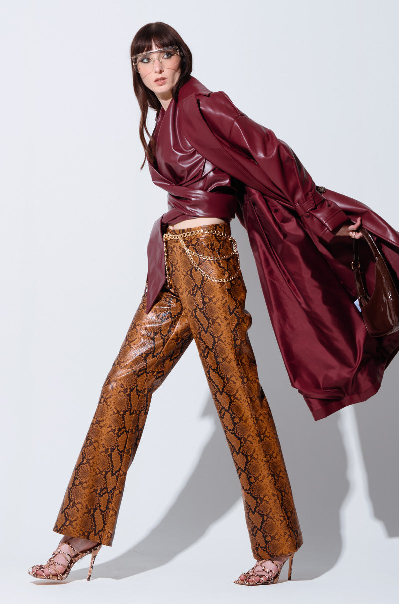 A woman with long dark hair poses dynamically against a plain background. She wears glasses, a deep burgundy coat, and snakeskin straight-leg trousers paired with matching high heels. Her coat billows behind her, adding a sense of movement to the image. 0