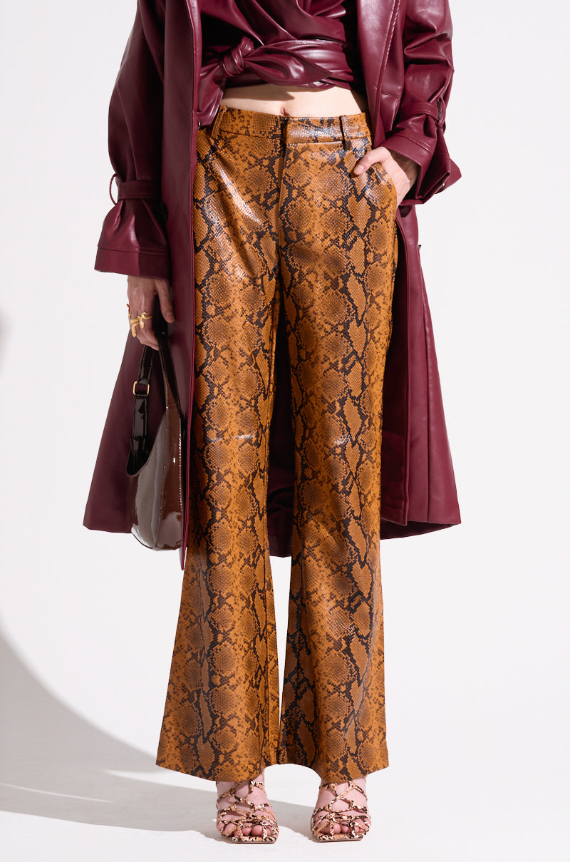 A person is wearing a burgundy coat and top, paired with SNAKESKIN STRAIGHT LEG TROUSER. They are also holding a dark handbag and wearing matching snake print sandals. The background is plain white. 1