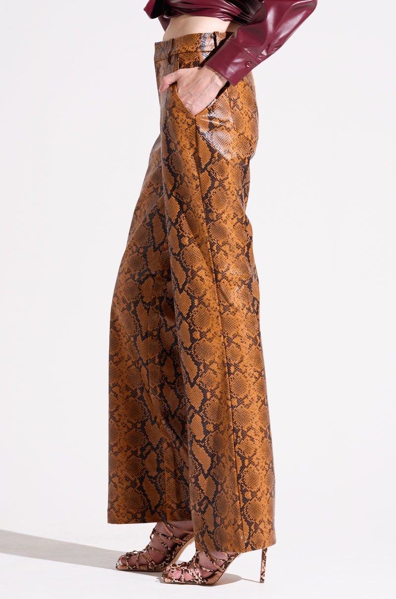 A person stands with one hand in their pocket, highlighting the SNAKESKIN STRAIGHT LEG TROUSERS adorned with a brown serpentine pattern. The outfit is complemented by high-heeled shoes featuring a matching snakeskin design. Only the lower half of the person's body is visible. 3