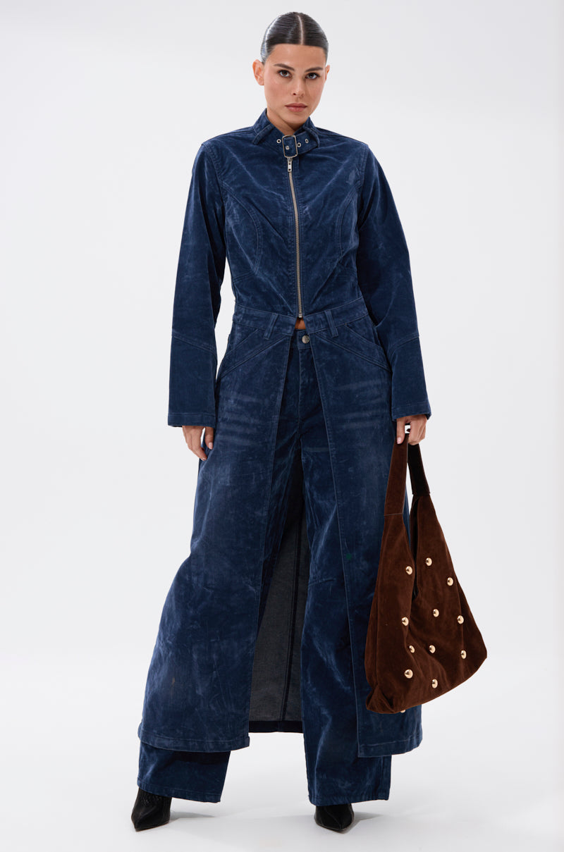A woman faces forward in the VALERIE VELVET EFFECT TRENCH IN BLUE, styled like wide-leg pants. She carries a brown suede bag with gold studs and wears her hair slicked back neatly.