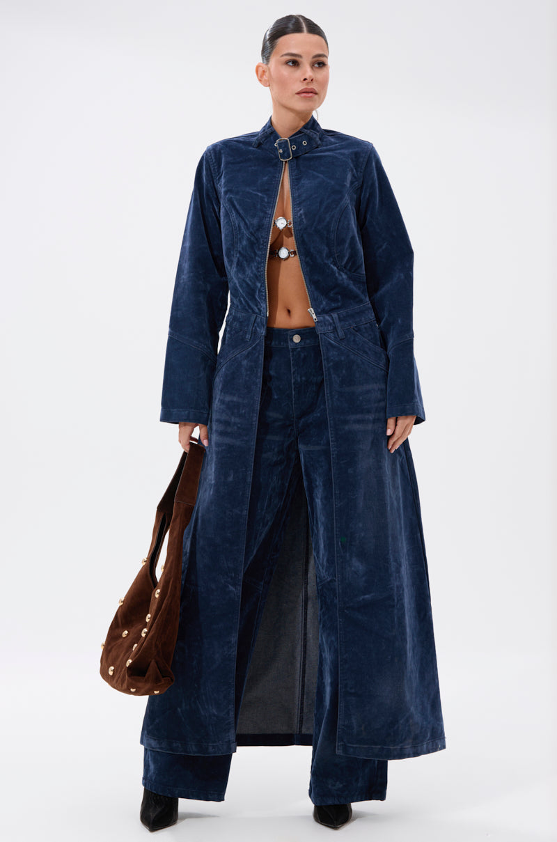 A woman in the VALERIE VELVET EFFECT TRENCH IN BLUE, paired with wide-leg pants and a revealing top, holds a brown handbag while standing against a plain white background.