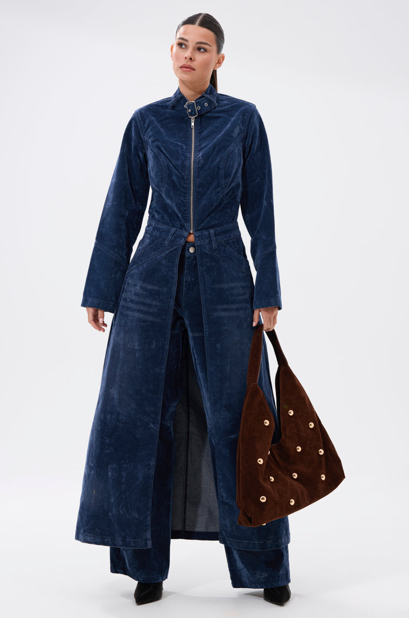 A woman wears the VALERIE VELVET EFFECT TRENCH IN BLUE with matching wide-leg pants and holds a brown suede shoulder bag with grommet details against a plain white background.