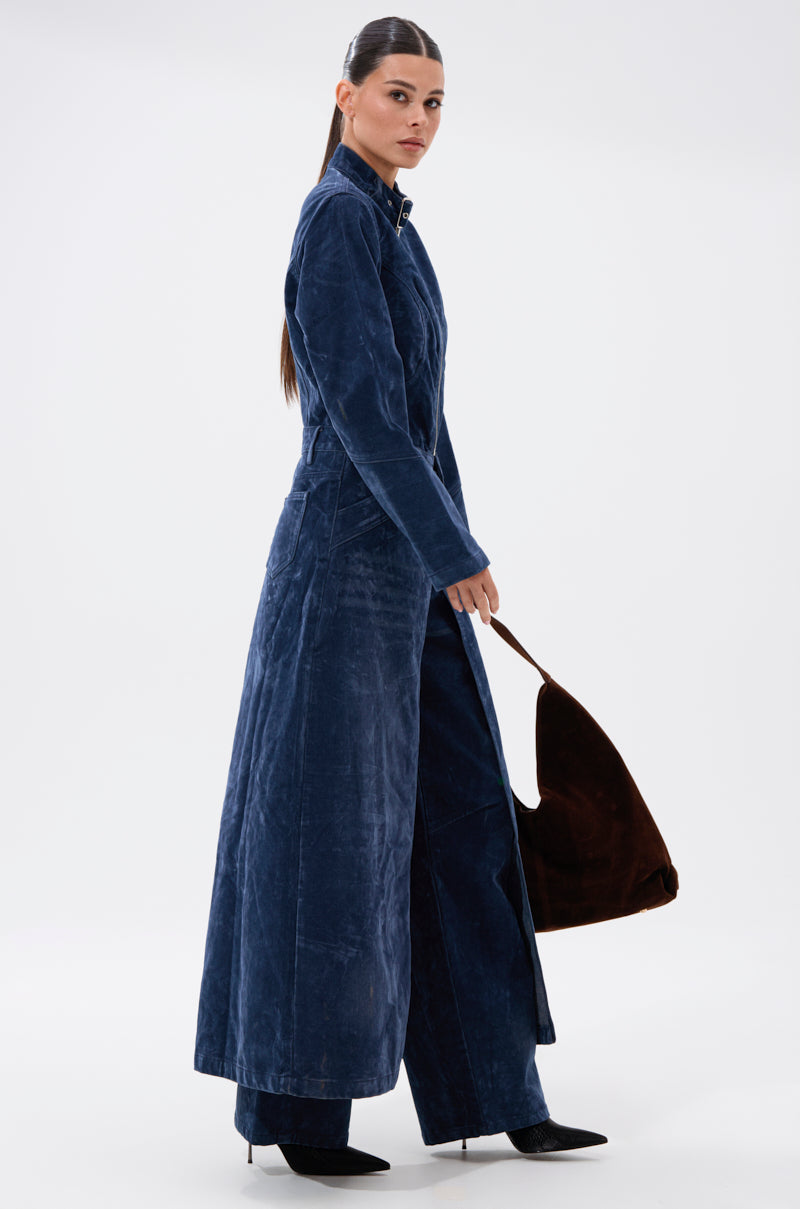A woman with long dark hair tied back wears the VALERIE VELVET EFFECT TRENCH IN BLUE over matching wide-leg pants, black pointed shoes, and holds a brown suede shoulder bag against a plain white background.
