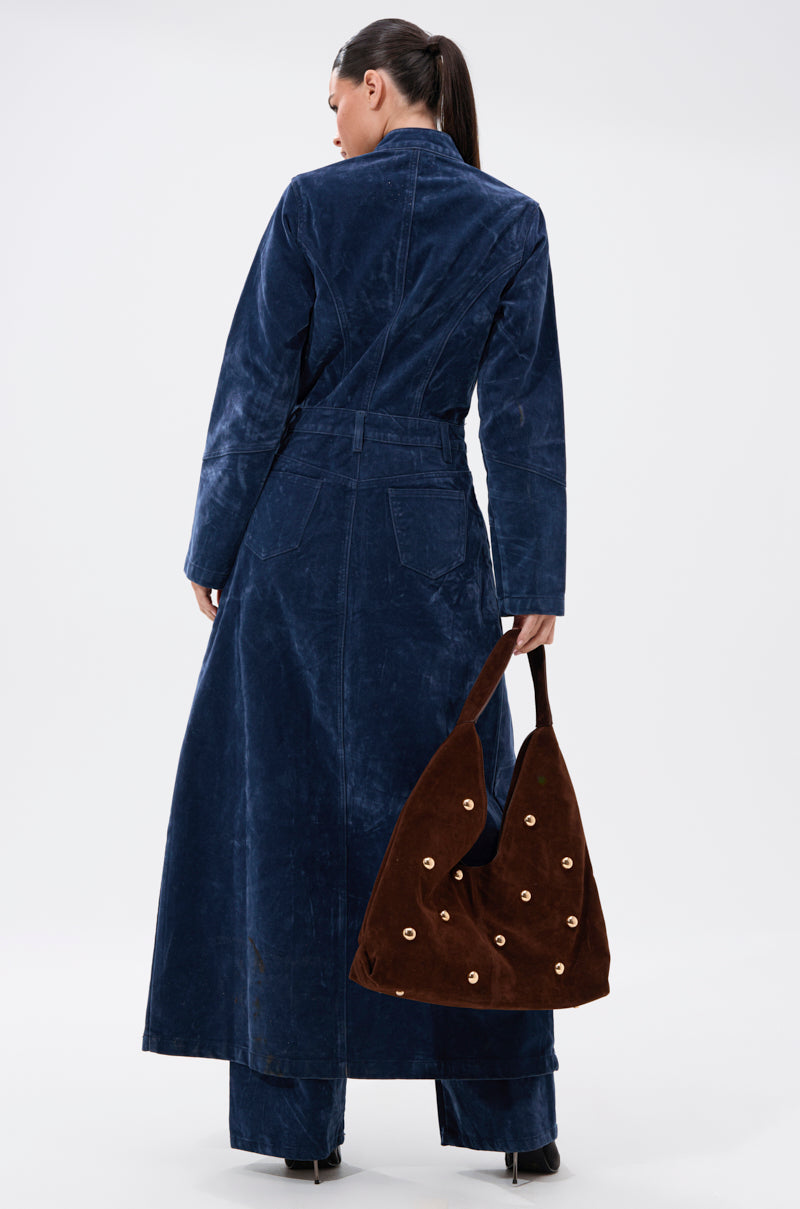 A woman with a ponytail, seen from behind, wears the VALERIE VELVET EFFECT TRENCH IN BLUE and matching pants. She carries a large brown suede shoulder bag with grommet details in her right hand against a white background.