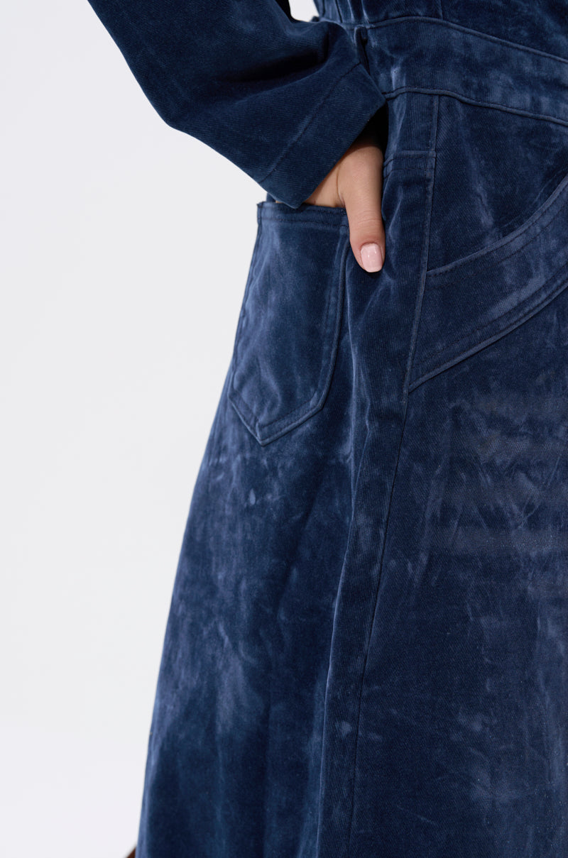 A close-up shows someone wearing the VALERIE VELVET EFFECT TRENCH IN BLUE, a dark blue velvet trench coat with a large pocket. Only their arm and torso are visible as their hand is partly tucked in, set against a light background.
