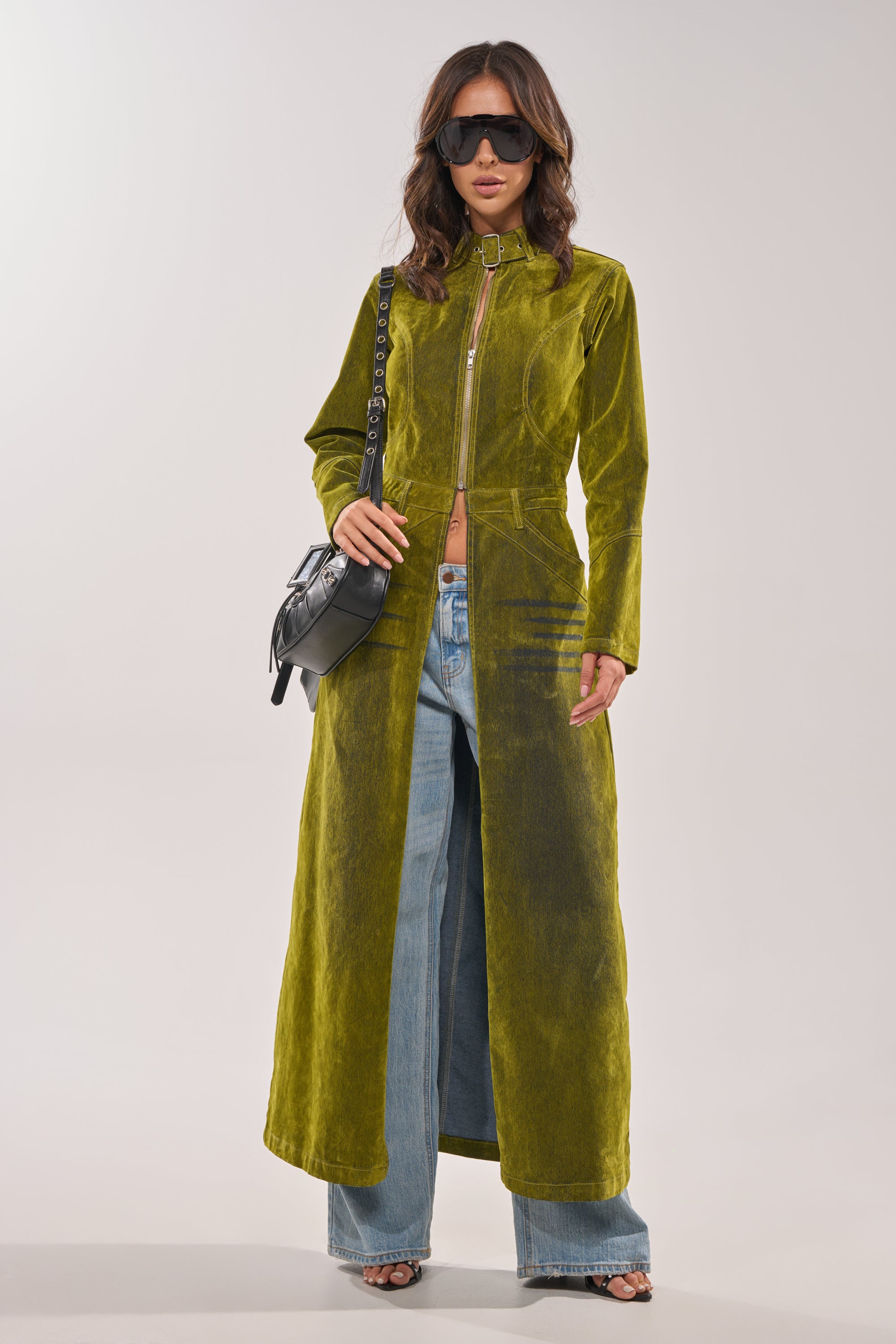 Wearing the VALERIE VELVET EFFECT TRENCH IN GREEN over blue wide-leg jeans, open-toed heels, sunglasses, and carrying a black shoulder bag, a woman stands against a plain light background. 2