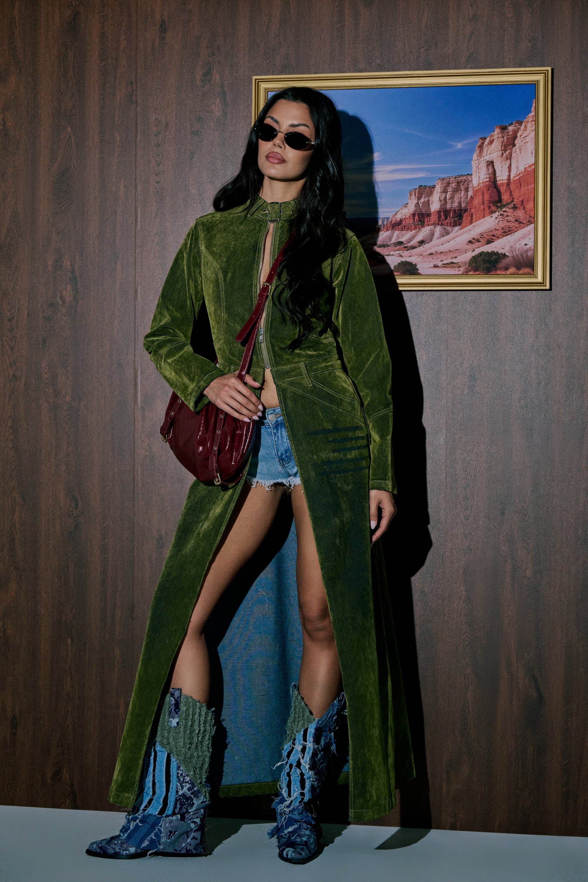 A woman poses indoors before a red rock cliff photo, wearing sunglasses, denim shorts, blue patterned boots, a maroon shoulder bag, and the VALERIE VELVET EFFECT TRENCH IN GREEN. 0