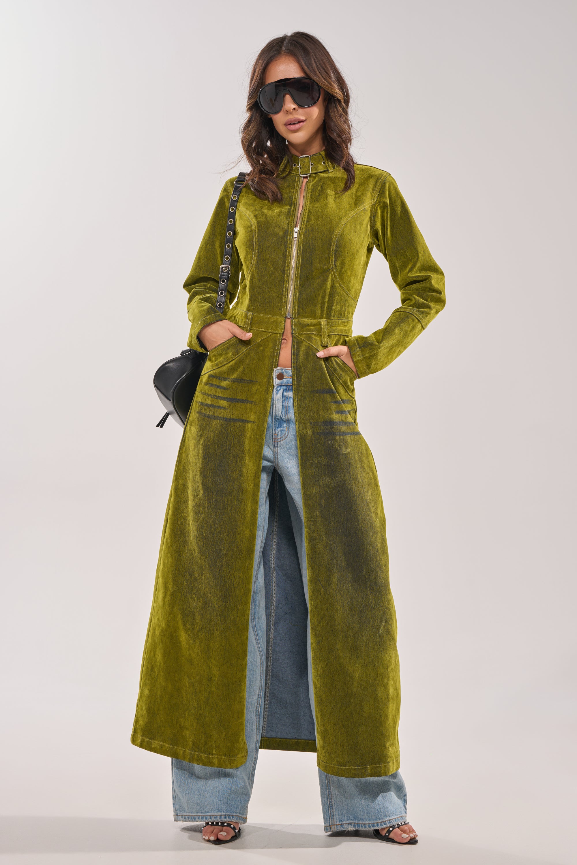 A woman poses confidently against a plain background wearing the VALERIE VELVET EFFECT TRENCH IN GREEN over wide-leg blue jeans, sunglasses, and holding a black chain-strap purse. 3