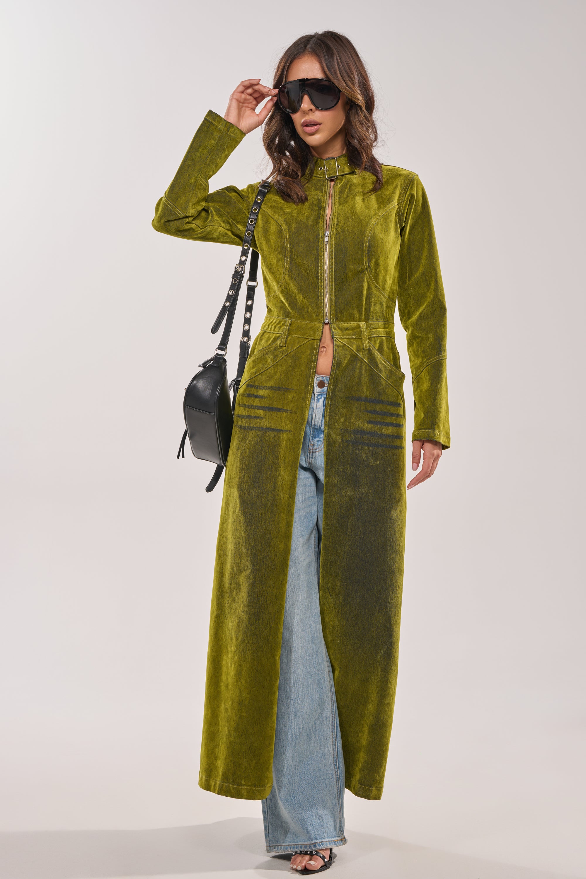 A woman in the VALERIE VELVET EFFECT TRENCH IN GREEN, styled over light blue jeans with black sunglasses and a small black handbag, stands against a plain light background. 4