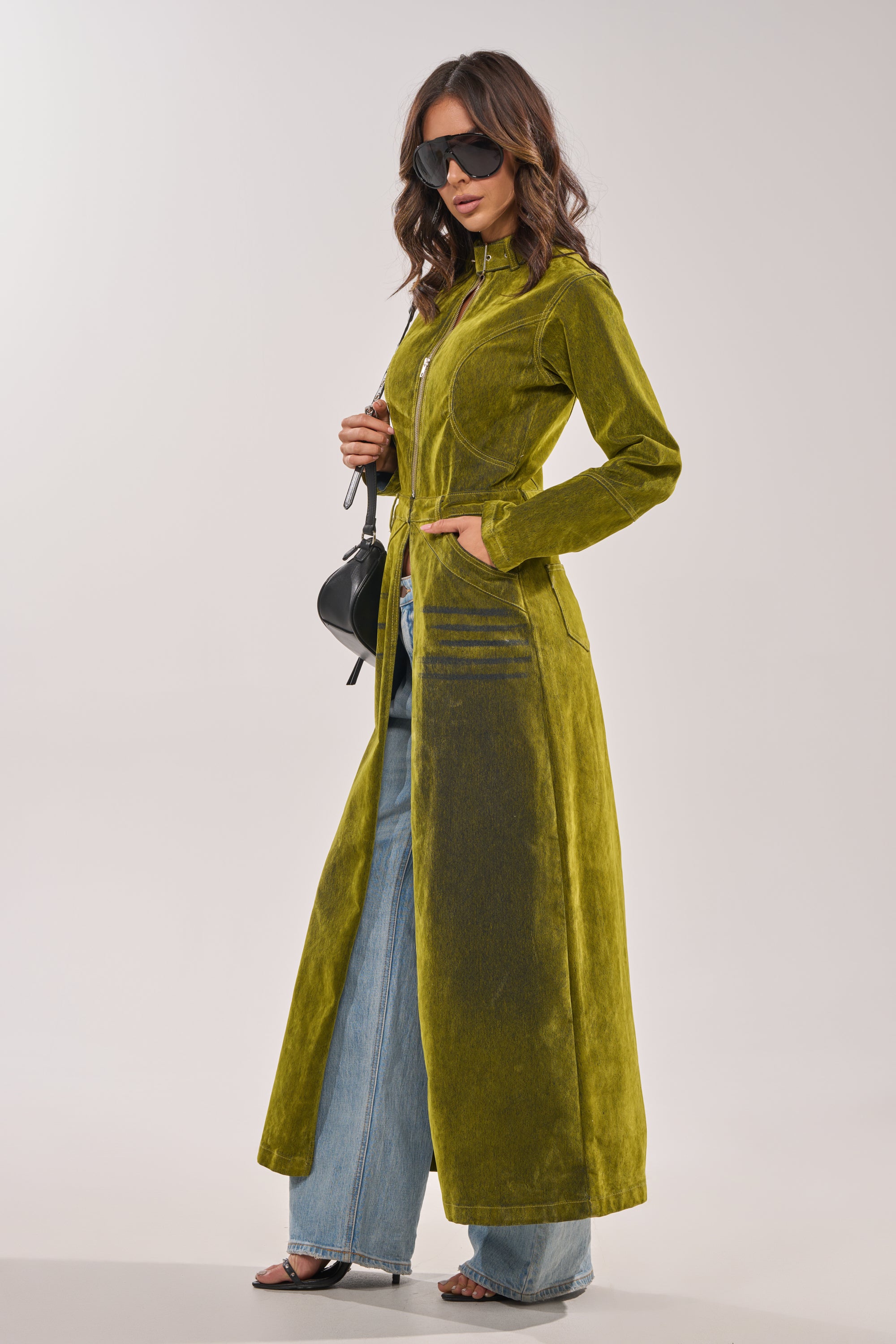 A woman poses against a neutral background wearing the VALERIE VELVET EFFECT TRENCH IN GREEN over wide-leg jeans and black heels, styled with large black sunglasses and a small black purse. 5