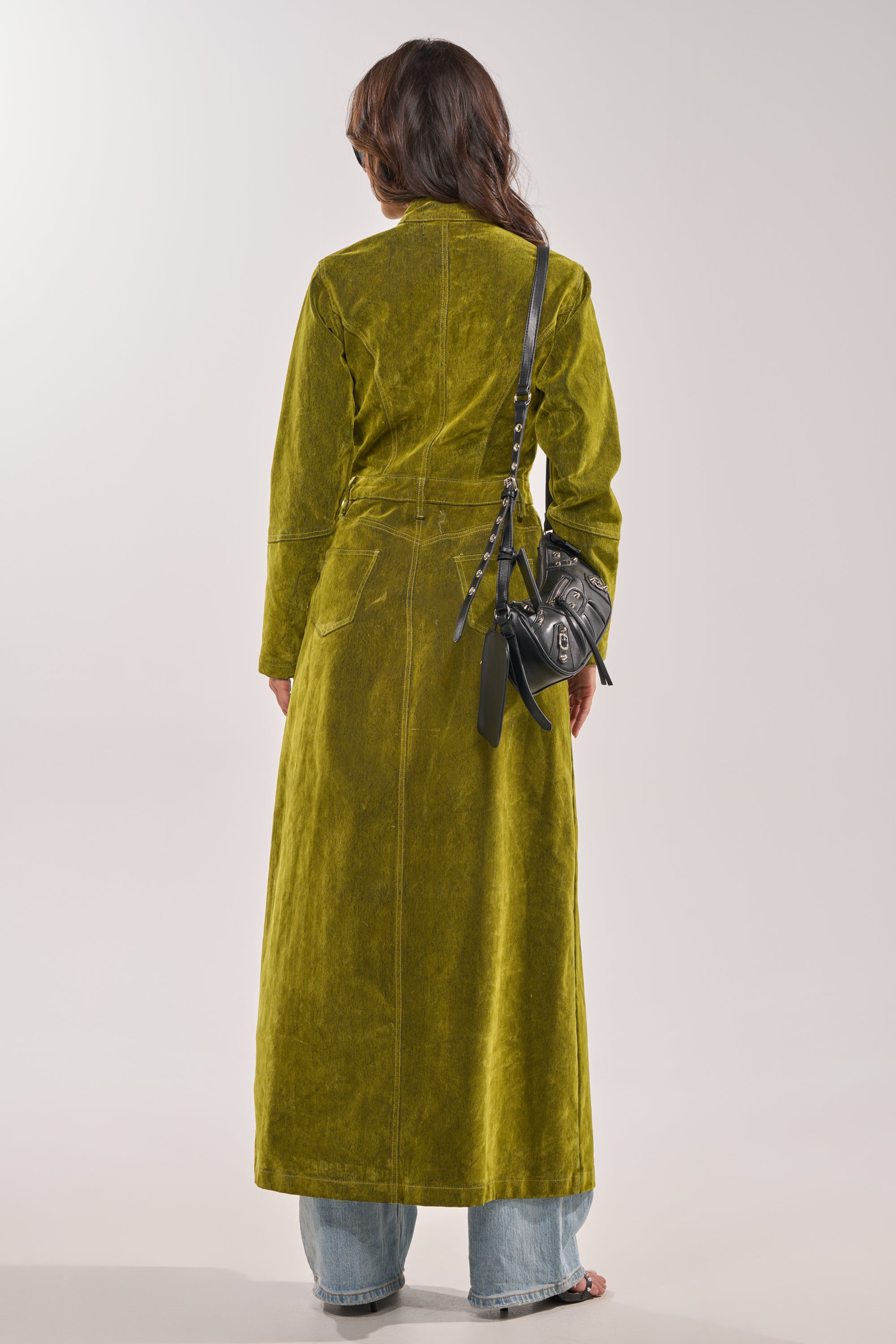 A person with wavy brown hair, wearing the VALERIE VELVET EFFECT TRENCH IN GREEN, light blue jeans, and a black crossbody bag stands facing away from the camera against a light background. 6