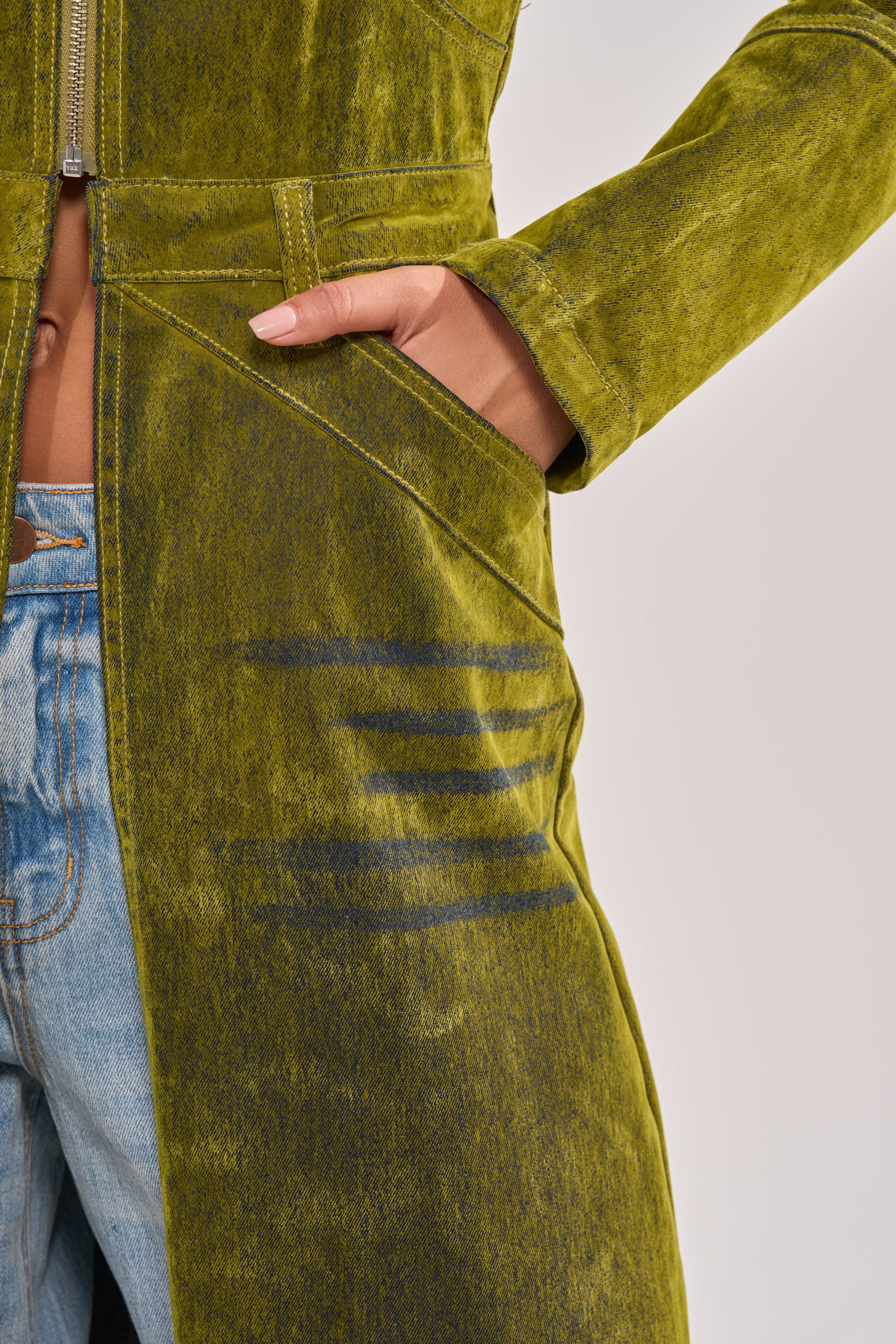 A person wearing blue jeans and the VALERIE VELVET EFFECT TRENCH IN GREEN with a hand in the pocket; the trench features dark horizontal streaks near the pocket and visible stitching details. 7