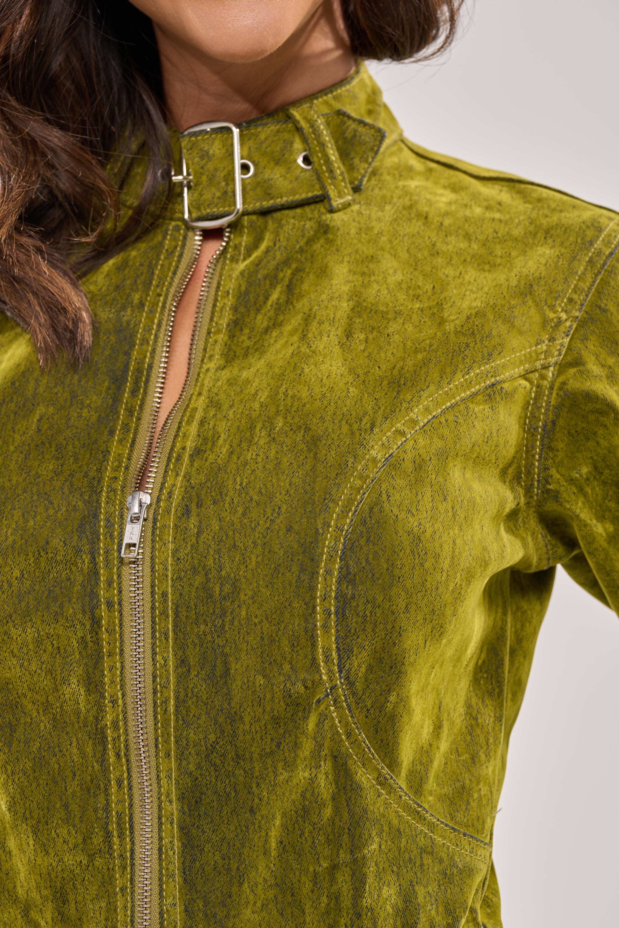 A close-up of a person wearing the VALERIE Velvet Effect Trench in green, featuring a longline fit, zipper and buckle collar, visible stitching, and round seam detail. Only the lower face and upper body are shown. 8