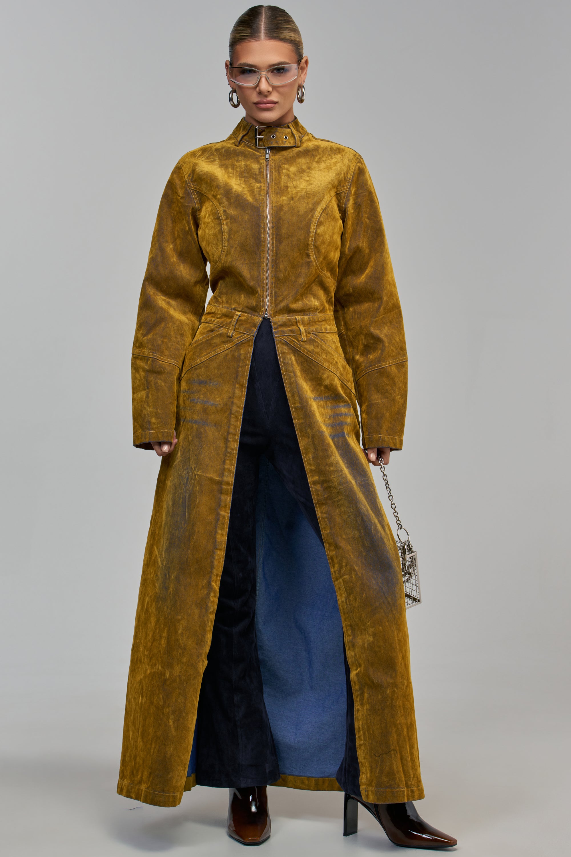 A model wearing a VALERIE VELVET DENIM TRENCH IN MUSTARD with a front zipper over dark trousers and brown heeled boots.