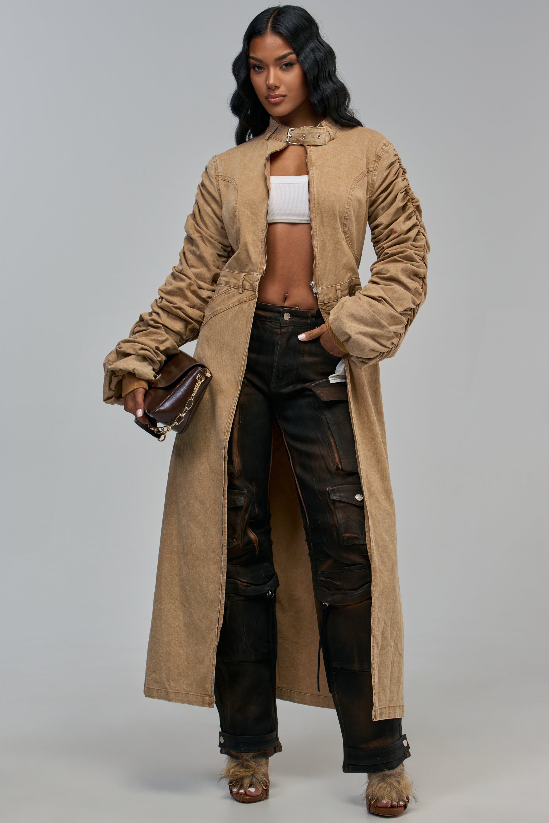 A woman wearing a JULES TRENCH IN KHAKI with ruched sleeves over a white crop top and dark, multi-pocketed cargo pants, holding a brown handbag.