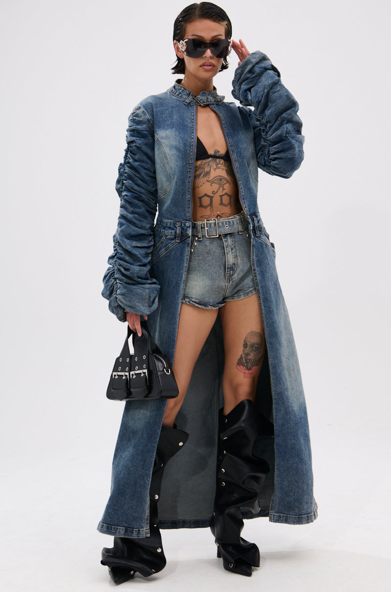 A model in the JULES DENIM TRENCH with ruched sleeves, denim shorts, black bra, futuristic sunglasses, knee-high black boots, and a small black handbag strikes a confident pose against a plain background. 1