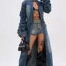 A model in the JULES DENIM TRENCH with ruched sleeves, denim shorts, black bra, futuristic sunglasses, knee-high black boots, and a small black handbag strikes a confident pose against a plain background.