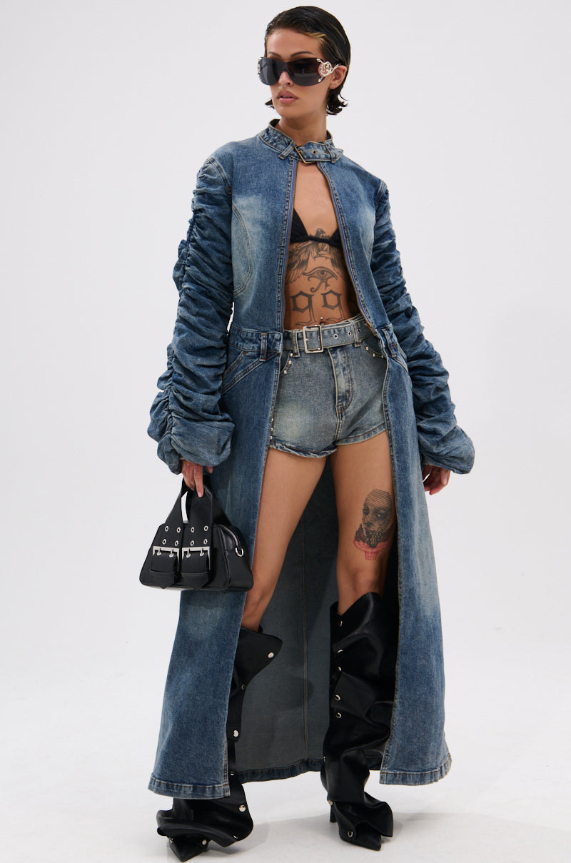 A person in oversized sunglasses, the JULES DENIM TRENCH with a longline silhouette, denim shorts, a black bikini top, tall black boots, and holding a small black handbag. Tattoos are visible on their torso and thigh. 2