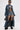 Wearing the JULES DENIM TRENCH in dark wash over a black bralette and denim shorts, paired with black platform boots, sunglasses, a choker necklace, and a small black handbag; tattoos accent legs and torso.