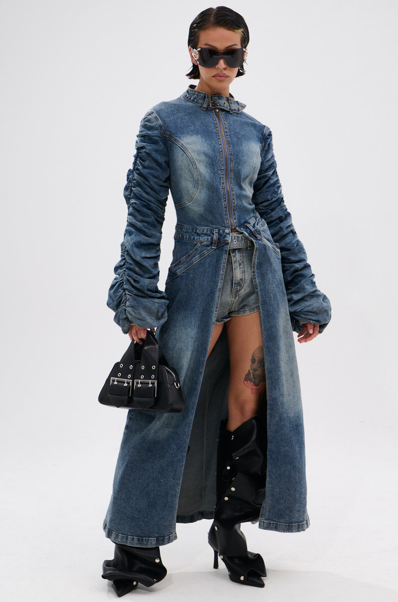 Wearing the JULES DENIM TRENCH with a longline silhouette over denim shorts, tall black heeled boots, and dark sunglasses, a person holds a small black handbag against a plain white background. 0