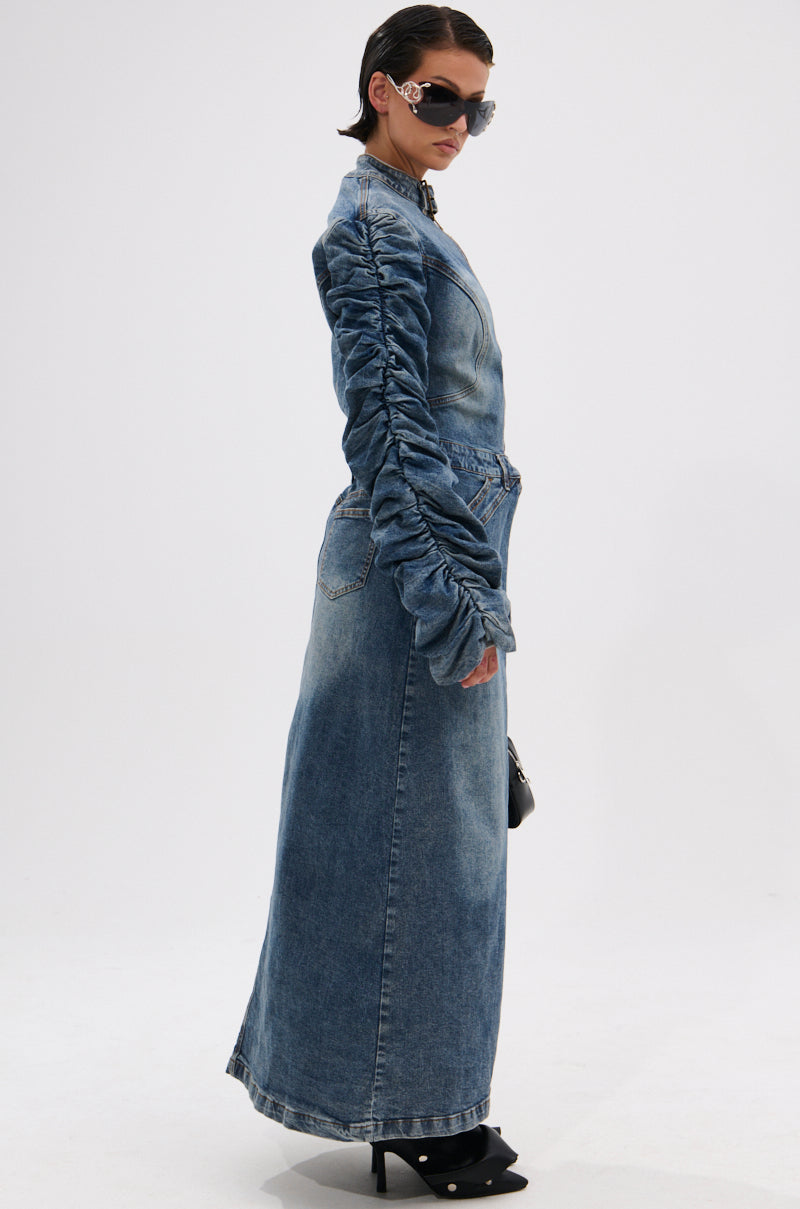 A person in profile wears the JULES DENIM TRENCH, a longline dark-wash denim coat with ruched sleeves, paired with a matching maxi skirt, black heels, sunglasses, and a small black bag. Their hair is slicked back; background is plain white. 4