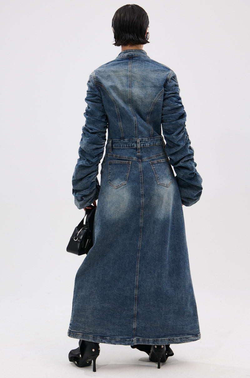 A person faces away wearing the JULES DENIM TRENCH, a dark-wash coat with ruched sleeves and a belted waist, styled with black heeled boots and a small black handbag against a plain white background. 5