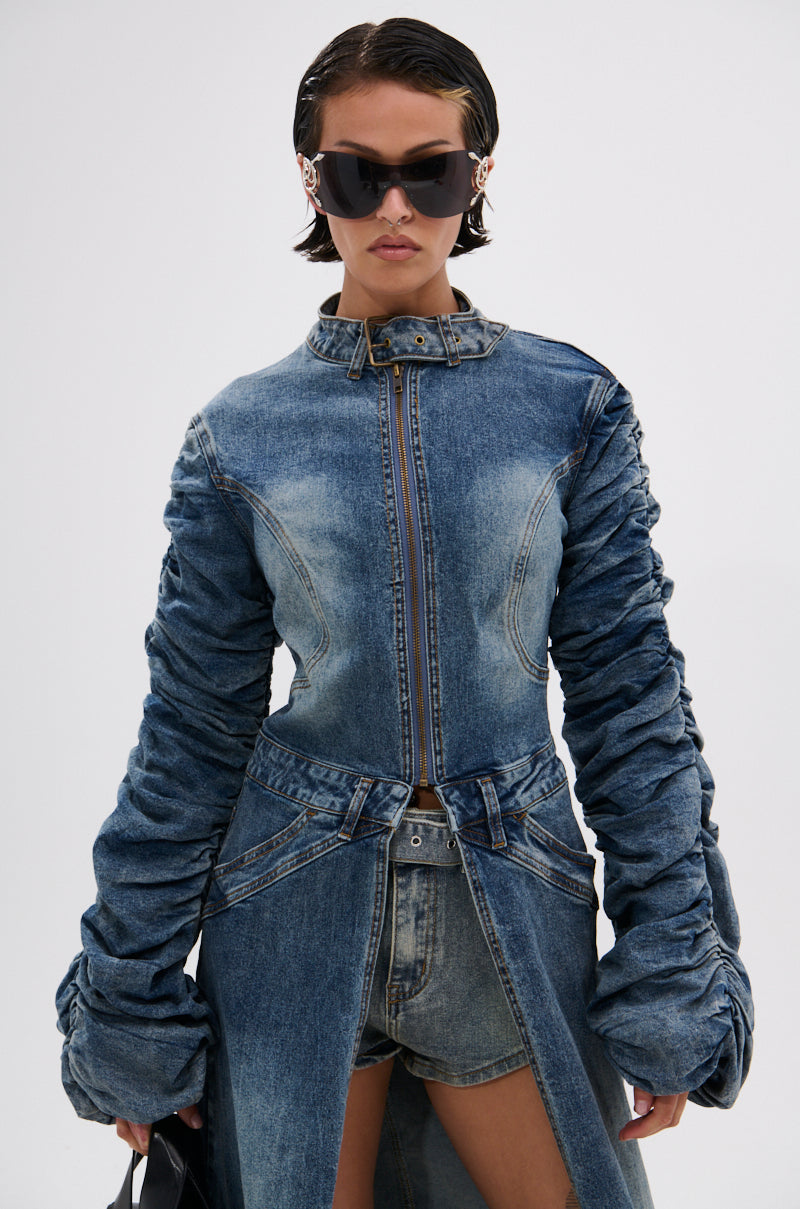 A woman with short, slicked-back hair stands against a white background wearing large black sunglasses, denim shorts, and the JULES DENIM TRENCH—a fitted dark-wash trench with ruched long sleeves. She carries a black bag. 6