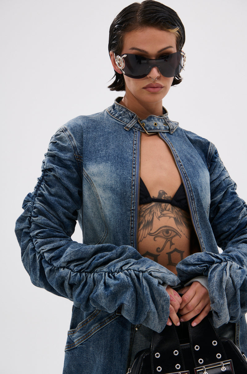 Wearing the JULES DENIM TRENCH in a dark wash, a person with short slicked-back hair, black bikini top showing a chest tattoo, large sunglasses, and studded black handbag stands against a plain light background. 7