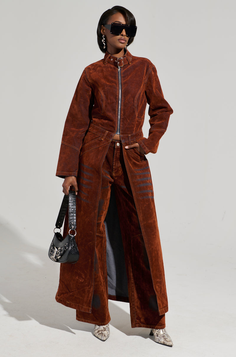 A person poses in the VALERIE VELVET EFFECT TRENCH, paired with wide-legged pants and a handbag. They wear oversized sunglasses and snakeskin-patterned shoes, exuding confidence against a plain background.